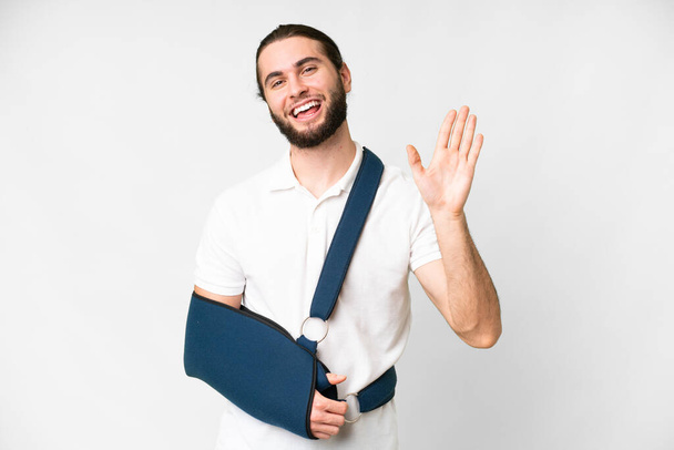 Young handsome man with broken arm and wearing a sling over isolated white background saluting with hand with happy expression - 写真・画像