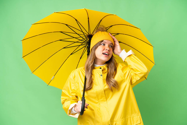 Młoda dziewczyna z płaszczem przeciwdeszczowym i parasolem na odizolowanym tle chroma key uśmiechając się dużo - Zdjęcie, obraz
