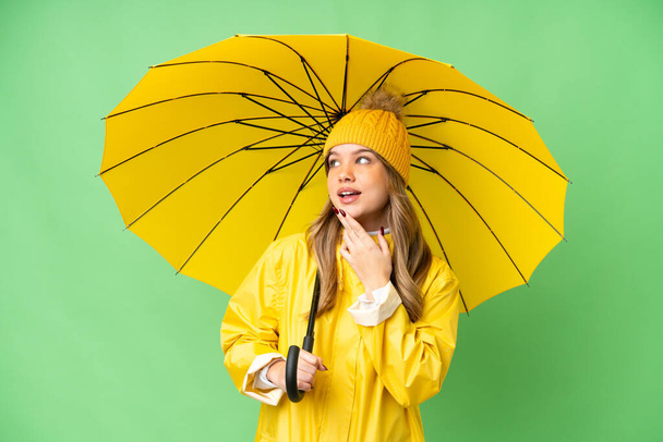 Młoda dziewczyna z płaszczem przeciwdeszczowym i parasolem na izolowanym tle chroma key patrząc w górę podczas uśmiechu - Zdjęcie, obraz