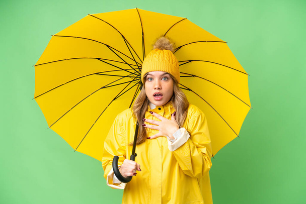 Yağmura dayanıklı ceketi ve şemsiyesi olan genç bir kız izole edilmiş krom anahtar arka planı üzerinde şaşırmış ve şaşırmış görünüyor. - Fotoğraf, Görsel