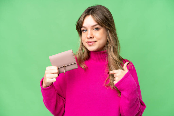Νεαρό κορίτσι κρατώντας ένα πορτοφόλι πάνω από απομονωμένο χρωματικό κλειδί φόντο με έκφραση έκπληξη προσώπου - Φωτογραφία, εικόνα