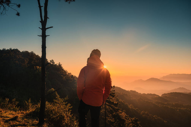 Οπίσθια όψη του ταξιδιώτη άνδρα φορώντας σακάκι στέκεται αντιπαράθεση με την ανατολή του ηλίου στην κορυφή του βουνού στο τροπικό δάσος στο εθνικό πάρκο - Φωτογραφία, εικόνα