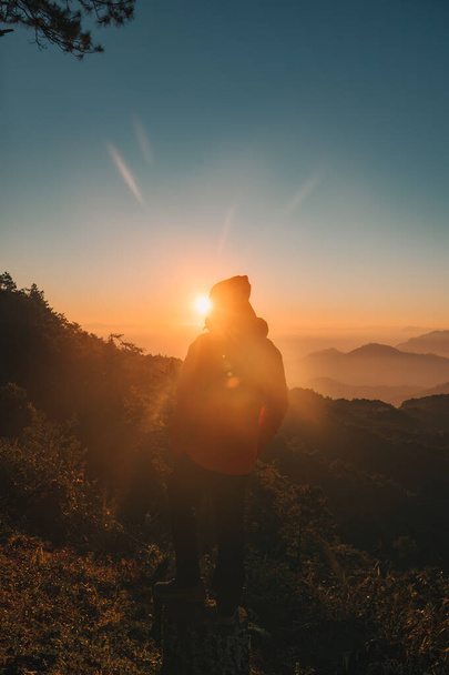 Вид сзади на человека в куртке, стоящего в противостоянии с восходом солнца на вершине горы в тропических джунглях национального парка - Фото, изображение