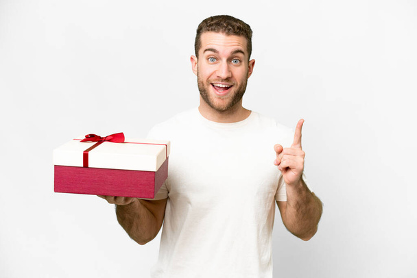 Fiatal jóképű szőke férfi kezében egy ajándék felett elszigetelt fehér háttér rámutatva egy nagy ötlet - Fotó, kép