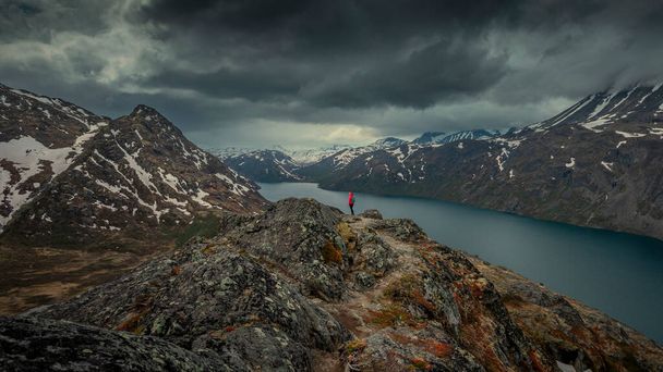 Женщина путешествует по голубому озеру в горном ландшафте Кнуттауг в Национальном парке Йотунхеймен в Норвегии, горы Бессегген на заднем плане, драматическое облачное небо - Фото, изображение