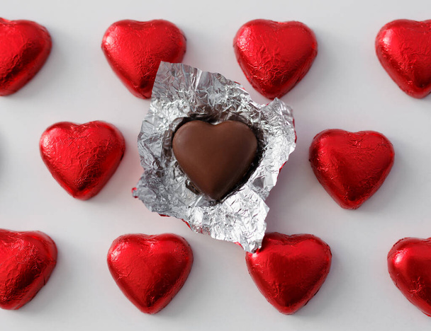 Love and relationship concept - close up of wrapped and unwrapped heart shape chocolate candies in red foil over white background - Zdjęcie, obraz