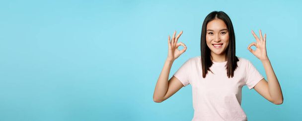 Geen probleem. glimlachende aziatische vrouw tonen oke tekenen, goedkeuring gebaar, blij en tevreden, staande over blauwe achtergrond. - Foto, afbeelding