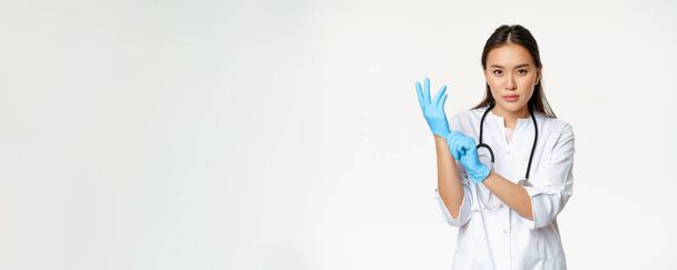 Γυναίκα νοσοκόμα με αυτοπεποίθηση, γιατρός που φοράει ελαστικά ιατρικά γάντια για την κλινική εξέταση του ασθενούς, στέκεται σοβαρή σε στολή του εργαζόμενου στον τομέα της υγειονομικής περίθαλψης, λευκό φόντο. - Φωτογραφία, εικόνα