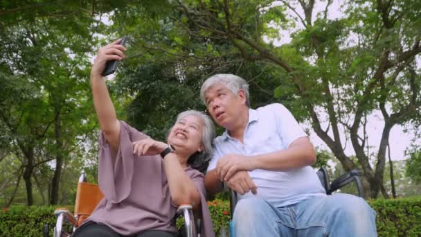 Šťastný asijské senior pár sedí na invalidním vozíku a používat smartphone fotit s manželem a mluvit spolu a relaxaci venku v parku, Volnočasové aktivity důchodců starší zdravotní péče - Záběry, video
