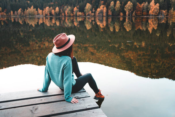 Side view of fashioned young woman sitting on wooden dock looking at view with beautiful autumn colors. Female hipster with brown hat relaxes on the edge of jetty. Wonderful nature getaway - Photo, Image