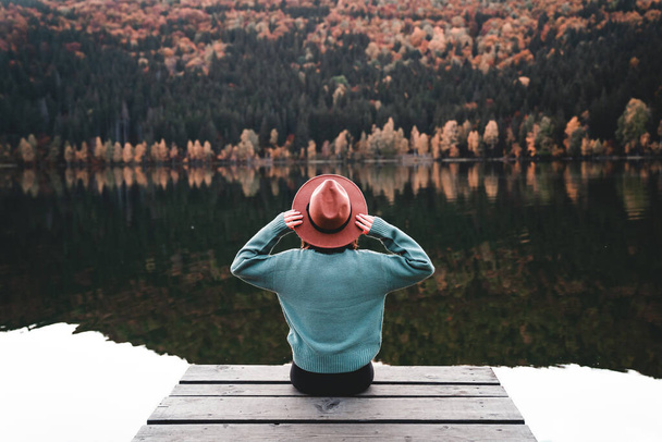 Back view of fashionable young woman sitting on wooden dock looking at view with beautiful autumn colors outdoors. Female hipster with brown hat relaxes on the edge of jetty. Wonderful nature getaway - Zdjęcie, obraz
