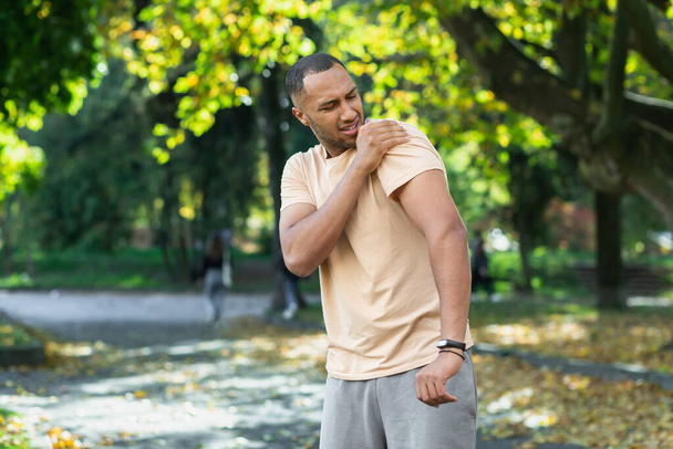 A man injured his shoulder during a fitness class, an African-American man injured himself while jogging in the park, stretches his arm and massages his sore muscles. - Foto, afbeelding