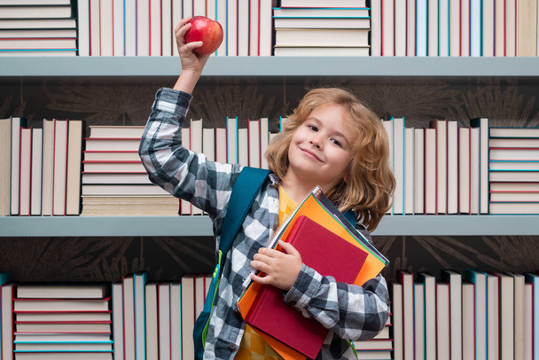 Kütüphanede kitapları ve elması olan okul çocuğu. Sevimli bir okul çocuğunun portresi. Okul ve eğitim çocukları konsepti. Okul malzemesi olan zeki bir çocuk. - Fotoğraf, Görsel