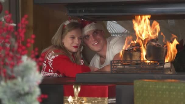 Loving Christmas couple leaning on each other and looking at burning fireplace. Cheerful man and woman wearing festive sweaters and enjoying romantic ambiance in home living room on Christmas Eve. - Filmagem, Vídeo