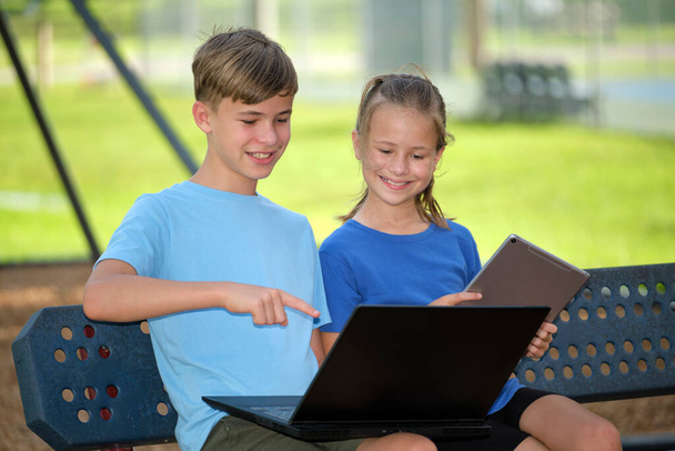 Dwójka dzieci, nastolatek trzymający laptopa i ładna młoda dziewczyna z cyfrowym tabletem ucząca się razem na zewnątrz, siedząca na ławce. Edukacja podczas kwarantanny. - Zdjęcie, obraz