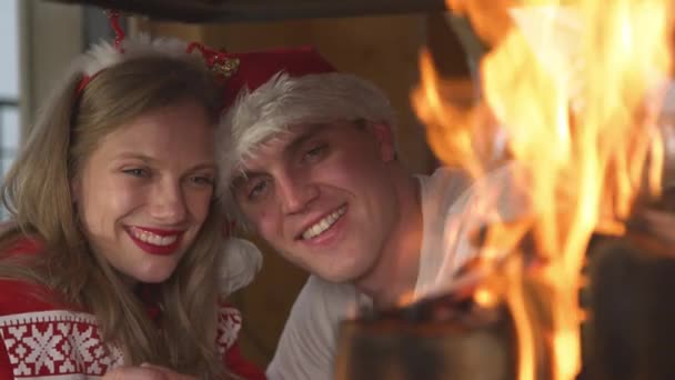 閉じる:ロマンチックなカップルがお互いに傾いて、燃える暖炉を見ています。笑顔の男と女性はお祝いのセーターを着て、クリスマスの夜に自宅のリビングルームでロマンチックな雰囲気を楽しんで. - 映像、動画