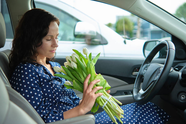 Porträt einer attraktiven jungen Frau in lässigem Kleid, die einen Blumenstrauß hält, während sie am Steuer eines Autos sitzt. - Foto, Bild