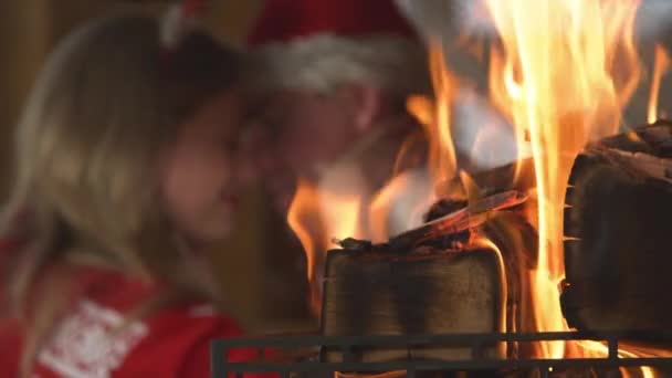 CLOSE UP: Burning fireplace and young couple in romantic mood in the background. View of the fire and young couple in love enjoying romantic atmosphere in home living room on festive winter Evening. - Felvétel, videó