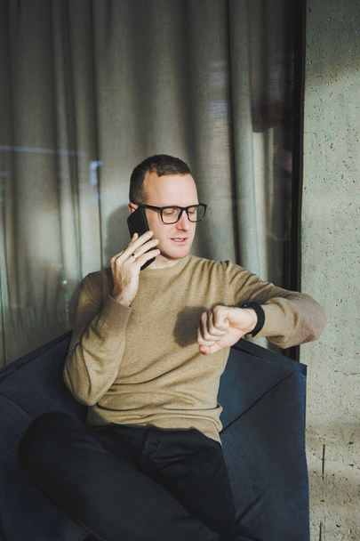A smiling young man in glasses is smiling and talking on the phone on a mobile phone while relaxing in the office. A young freelancer works remotely. - Foto, Bild