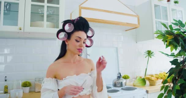Seksikäs 35-vuotias ruskeaverikkö nainen valkoisissa alusvaatteissa seisoo keittiössä ja lisää hiusneulan hiuksiinsa. Pystysuora 4k-kuvamateriaali. - Materiaali, video