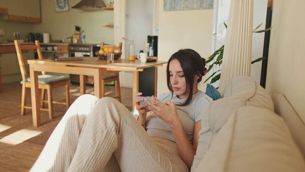 Young woman using mobile phone and changing channels from remote control while sitting on sofa at home - Foto, afbeelding