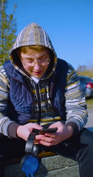 Концепция пристрастия к смартфонам - крупный план подростка со смартфоном и наушниками - он беседует с друзьями на открытом воздухе. Съемки крупным планом по вертикали. - Кадры, видео