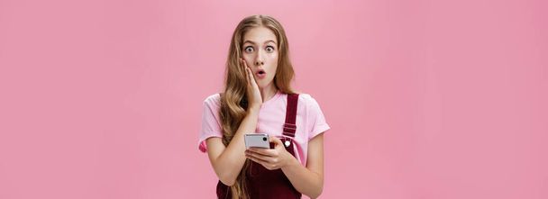 Vyötärö-up laukaus yllättynyt hämmästynyt järkyttynyt nuori hoikka nainen aaltoileva hiukset haalarit koskettaa poskea järkytys tilalla älypuhelin reagoi vaikuttava odottamattomia uutisia vaaleanpunainen seinä. Teknologia - Valokuva, kuva