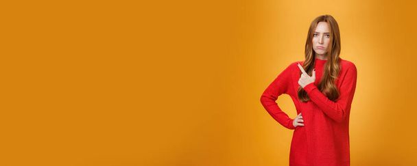 Rozrušený a zklamaný mladý zrzavý dívka v červeném svetru ukazuje na levém horním rohu nebo za zamračený vypadající zachmuřený a naštvaný, stojí žárlivý a náladový na oranžovém pozadí. - Fotografie, Obrázek
