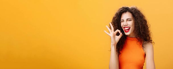 Stylish self-assured caucasian girlfriend. with curly hairstyle winking joyfully and sticking out tongue showing perfect or okay gesture assuring she got everything under control over orange wall. - Photo, Image