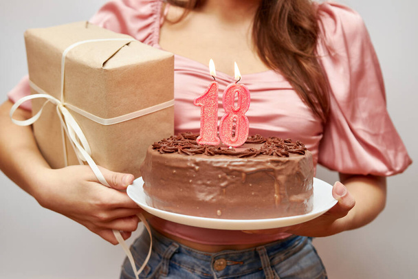 Het meisje houdt een feestelijke taart met een kaars in de vorm van het nummer 18 en een geschenk. Verjaardagsviering concept. - Foto, afbeelding