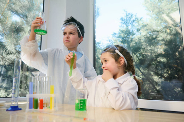 İki zeki çocuk, genç bir oğlan ve kreş kızı, beyaz laboratuvar önlüğü giyiyorlar, okul fen laboratuarında kimyasal deneyler yapıyorlar. Masada çeşitli kimyasallar ve maddeler var. - Fotoğraf, Görsel