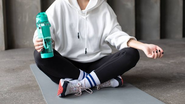 The athlete is resting after training, sitting on the mat and holding a reusable sports bottle with water. Healthy lifestyle concept, reusable bottle. Copy space - Photo, image
