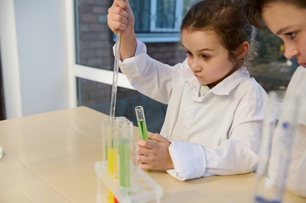 Mischievous Caucasian 5 years old little child girl in white lab coat, conducting chemical experiments, dripping few reagents into a test tube in the chemistry class. Back to school concept - Foto, immagini