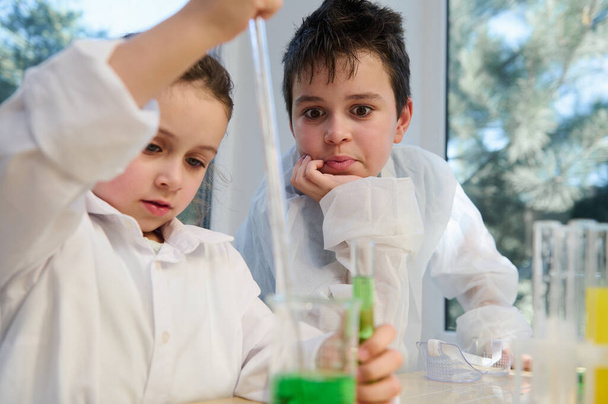 Кавказский умный подросток в белом халате, наблюдает за химической реакцией, происходящей в пробирке и стакане, проводя с одноклассником эксперимент в лаборатории на уроке химии - Фото, изображение