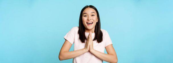 Enthousiaste Aziatische vrouw kijkt hoopvol, zeg alsjeblieft, tonen bedelend gebaar, vragen om smth, staan over blauwe achtergrond. - Foto, afbeelding