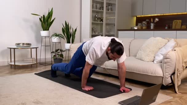 Sporty man working out following online training guidance on laptop at home. Mid adult bearded male doing bear crawl exercise watching video tutorial indoors. Athletic guy exercising in domestic room - Felvétel, videó