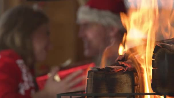 CLOSE UP: Lit fireplace and man giving a woman Christmas present in background. View of fireplace and young twosome celebrating festive winter Evening and enjoying cosy atmosphere in home living room - Materiał filmowy, wideo