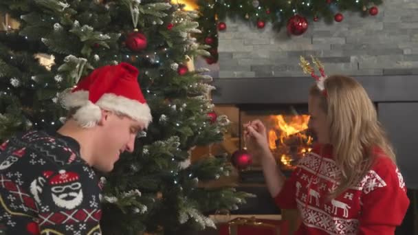 暖炉でクリスマスツリーを飾るお祝いの衣装でかわいいカップル。お祝いの冬の休日を祝うために自宅のリビングルームでクリスマスの装飾を準備する若い男と女. - 映像、動画
