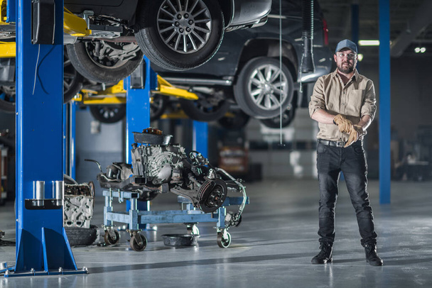 Caucasian Mechanic Getting Ready to Install the Drive Train. Professional Automobile Repair Workshop with Vehicles Secured on Car Lifts in the Background. - Fotoğraf, Görsel