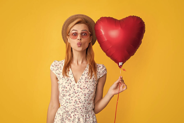 positive young woman in straw hat and sunglasses hold love heart balloon on yellow background. - 写真・画像
