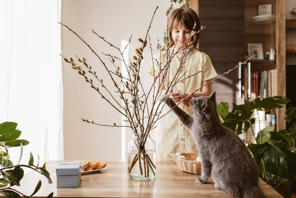 A happy girl hangs Easter eggs on a willow bouquet at home with a little kitten. Funny pets. Easter decoration. - Foto, immagini