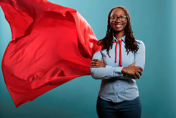 Brave and happy looking young adult superhero woman wearing red hero cape while smiling at camera. Justice defender with superpowers and mighty posture standing with arms crossed on blue background. - Photo, Image