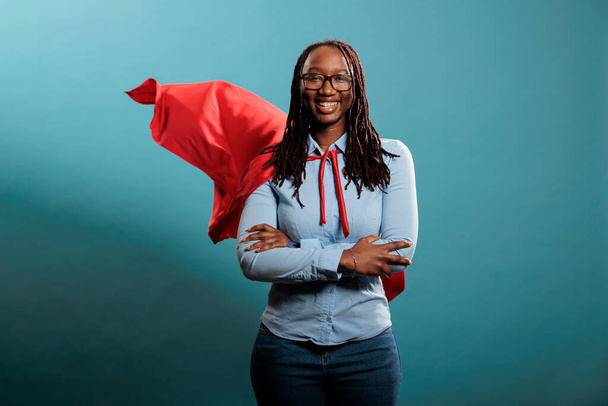 Confident looking mighty powerful young adult person wearing superhero costume on blue background. Portrait of happy brave woman with superpowers wearing red hero cape while standing with arms crossed - Foto, imagen