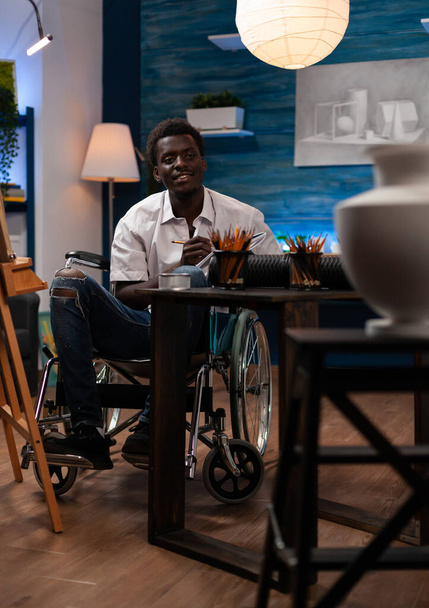Африканский американец делает набросок керамической вазы в домашней художественной студии. Художник-инвалид-колясочник с помощью карандаша и бумаги создает шедевр скульптуры в спальне. - Фото, изображение
