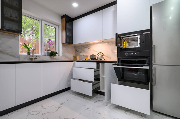 A contemporary kitchen with white cabinets, a marble floor, an open oven door, and pull-out shelves for easy access to ingredients and appliances. - Foto, Imagen