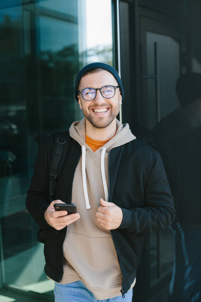 Позитивный молодой парень держит в руках смартфон, смотрит в камеру, стоящую на улице. Бородач в очках носит повседневную одежду. Концепция использования мобильных телефонов - Фото, изображение