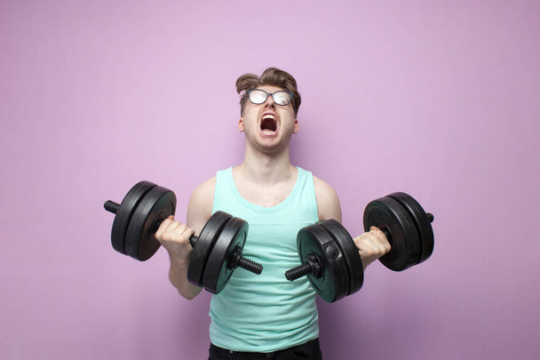 νεαρός μη-αθλητικός τύπος με γυαλιά κρατά βαριά αλτήρες σε ροζ φόντο και τρένα, ένα σοβαρό κίνητρο σπασίκλας πηγαίνει για σπορ και ουρλιάζοντας - Φωτογραφία, εικόνα