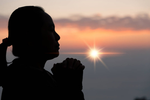 Faith of christian concept. Spiritual prayer hands over sun shine with blurred beautiful sunset background. Female hands worship God with love and faith. silhouette of a woman praying with a cross. - Photo, image