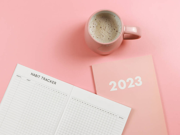 Bovenaanzicht of platte lay-of gewoonte tracker boek op roze dagboek of planner 2023 en roze kopje koffie op roze achtergrond met kopieerruimte. - Foto, afbeelding
