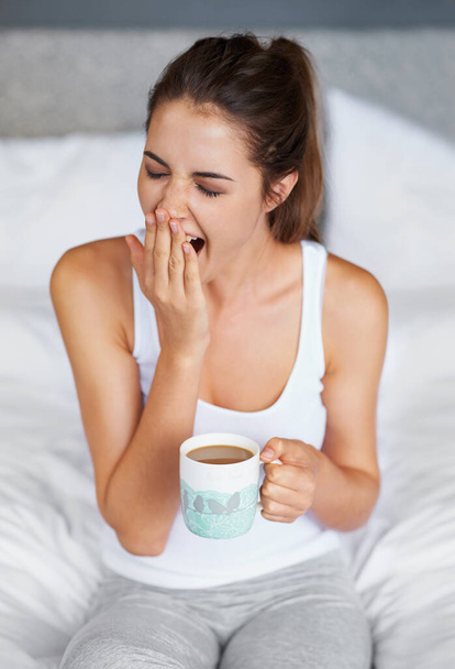 まだとても眠い...コーヒーを飲みながらベッドに座っている間に大きなあくびをする若い女性 - 写真・画像
